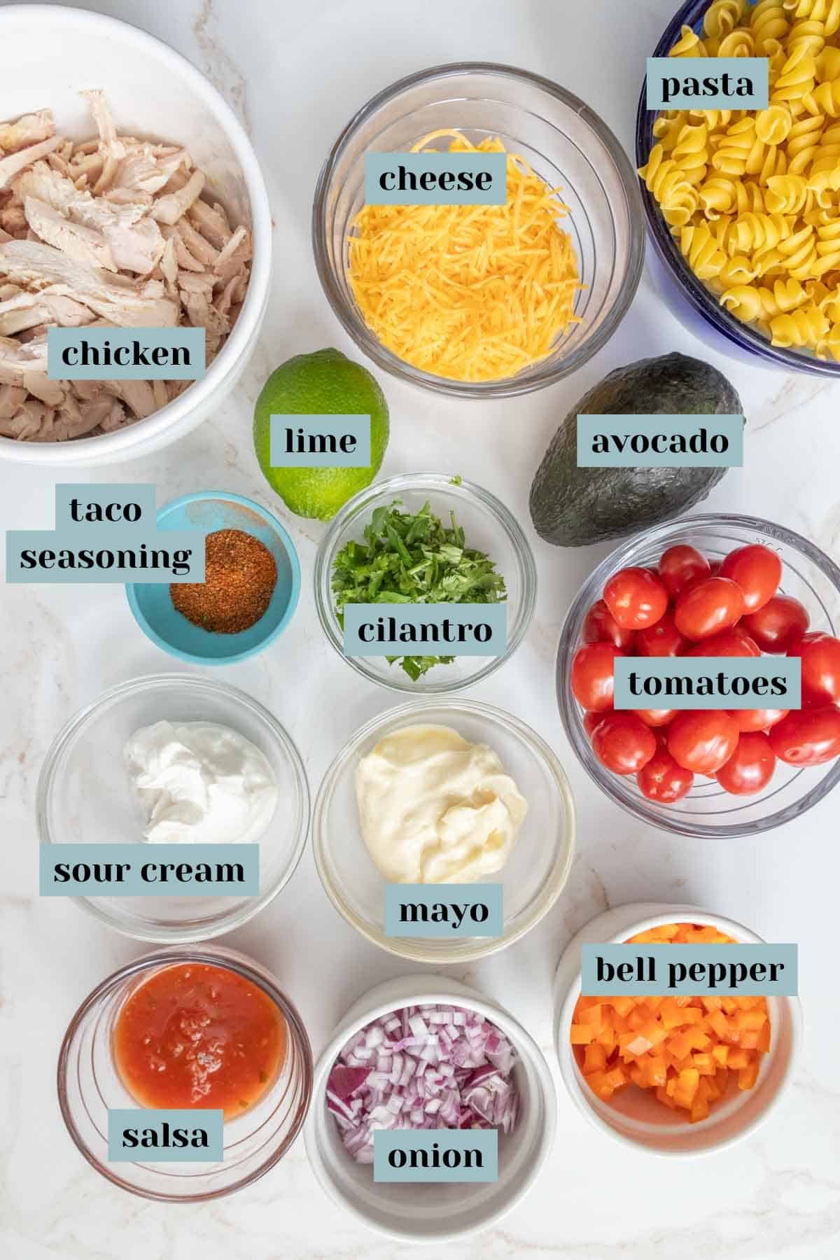 Various ingredients for a meal are laid out in bowls on a countertop, including pasta, shredded chicken, cheese, lime, avocado, taco seasoning, cilantro, tomatoes, sour cream, mayo, salsa, onion, and bell pepper.