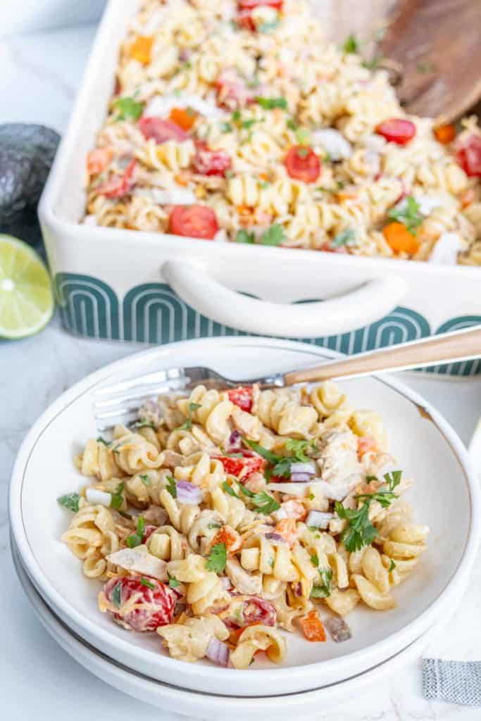 A white bowl filled with pasta salad containing rotini pasta, diced tomatoes, red onions, and parsley. A larger casserole dish with additional pasta salad is in the background. An avocado and lime are nearby.