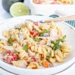 A bowl of creamy pasta salad with rotini, cherry tomatoes, red onions, bell peppers, and cilantro, garnished with a lime slice. An avocado and a baking dish of the pasta salad are in the background.