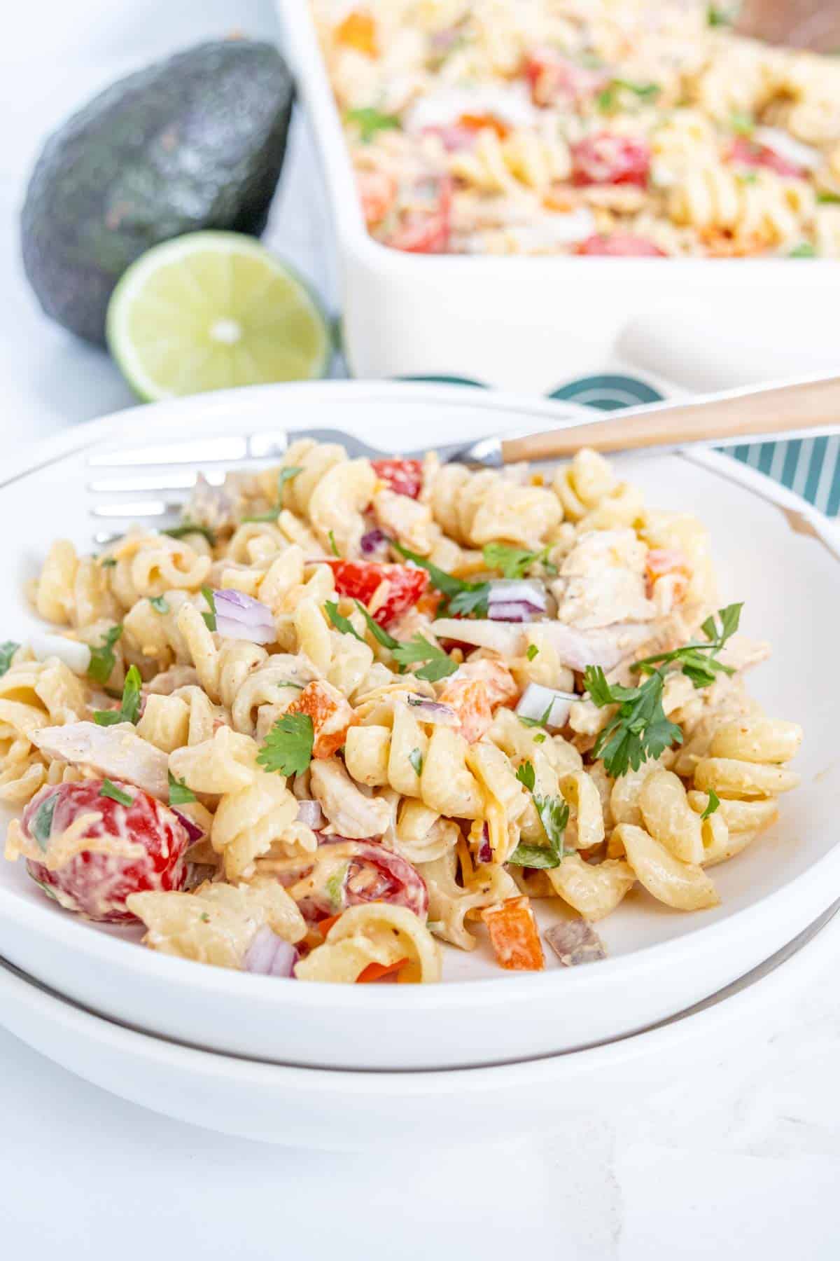 A bowl of creamy pasta salad with rotini, cherry tomatoes, red onions, bell peppers, and cilantro, garnished with a lime slice. An avocado and a baking dish of the pasta salad are in the background.