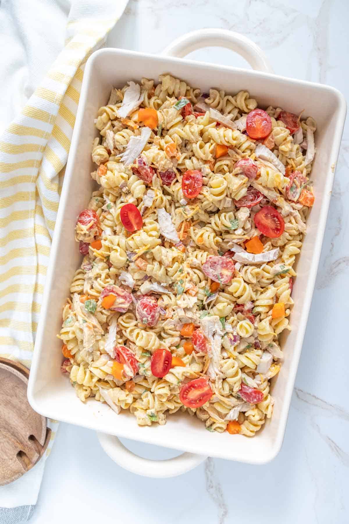 A rectangular dish of rotini pasta salad with sliced cherry tomatoes, shredded chicken, and a variety of chopped vegetables on a marble surface. A yellow and white striped cloth is beside the dish.