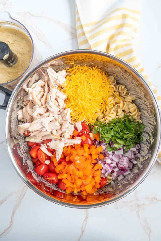 A large metal bowl contains shredded chicken, grated cheese, rotini pasta, chopped tomatoes, diced bell peppers, red onion, and cilantro. A food processor and a striped cloth are in the background.