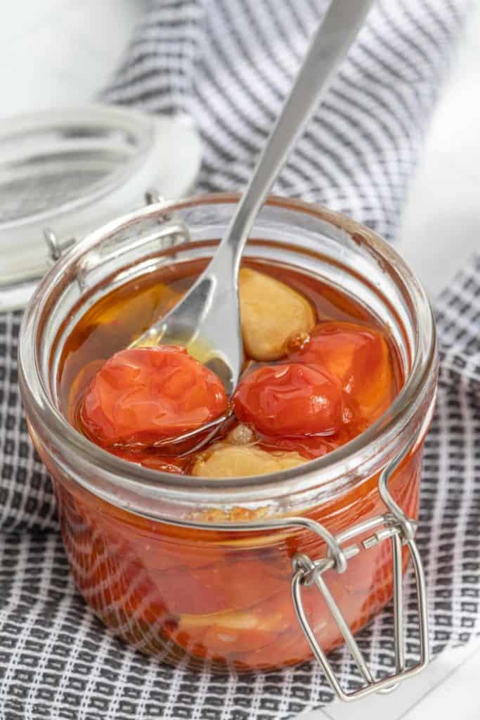 A glass jar filled with cherry tomatoes and garlic in oil, with a spoon placed inside. The jar is placed on a black and white checkered cloth. The lid of the jar is open and rests beside it.