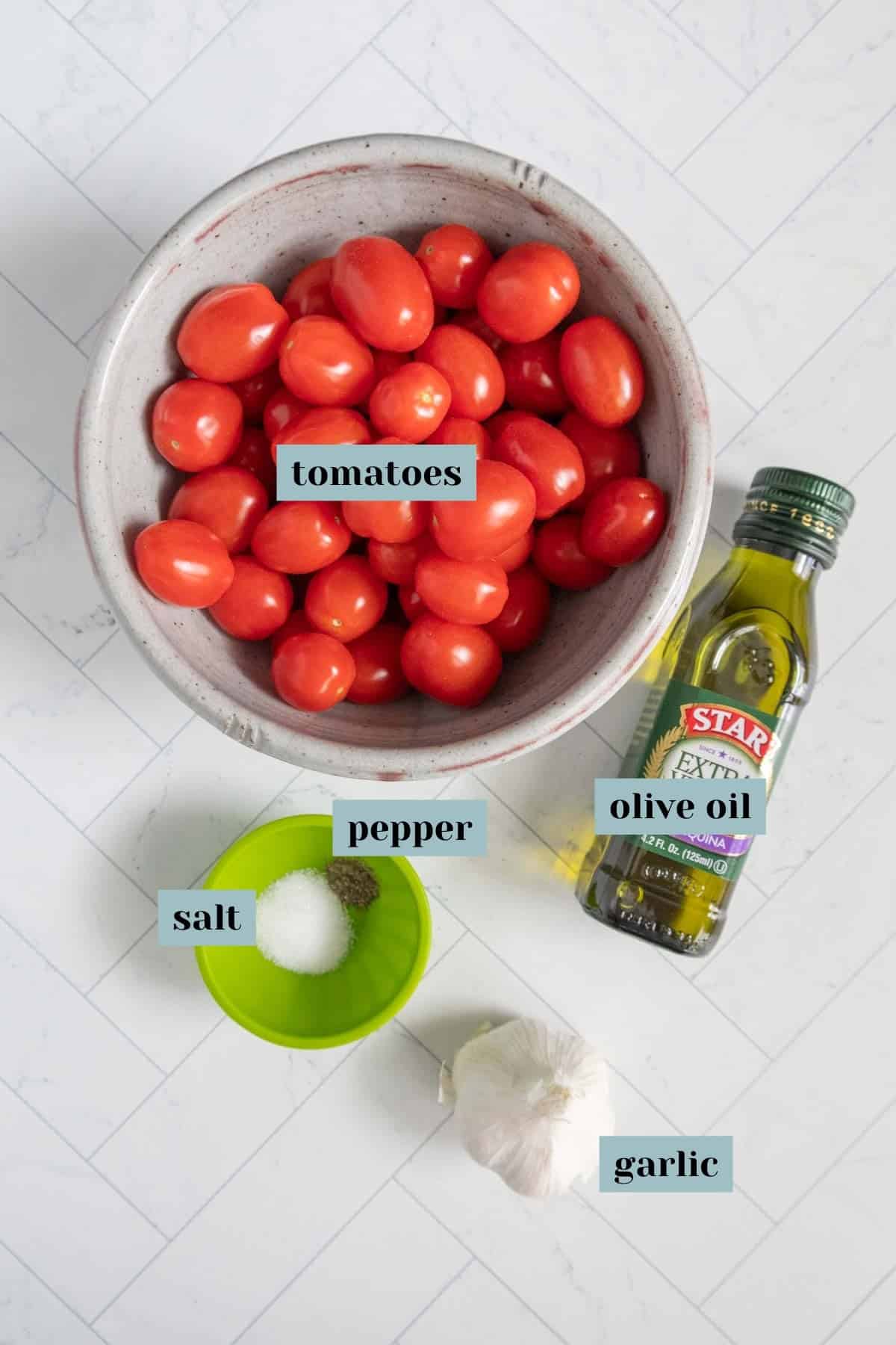 A bowl of tomatoes, a bottle of olive oil, a green dish with salt and pepper, and a bulb of garlic arranged on a white tiled surface.