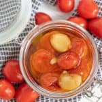 A glass jar filled with tomatoes and garlic cloves in oil, placed on a black-and-white checkered cloth, surrounded by whole tomatoes.