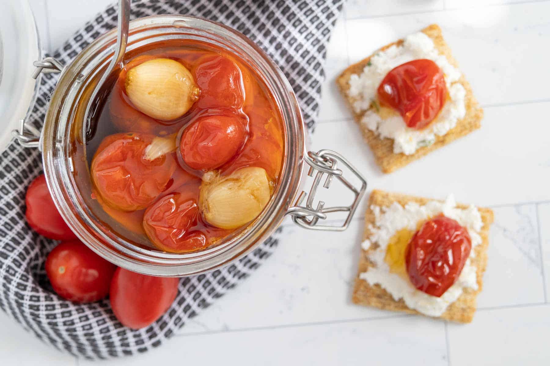 Open jar of marinated cherry tomatoes and pearl onions on a checkered cloth, next to a few fresh tomatoes. Two crackers topped with cheese and cherry tomatoes are on the side on a white surface.