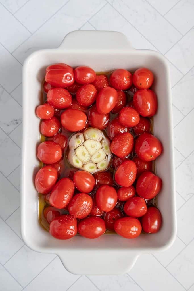 A white baking dish filled with cherry tomatoes, olive oil, and a halved garlic bulb on a tiled surface.