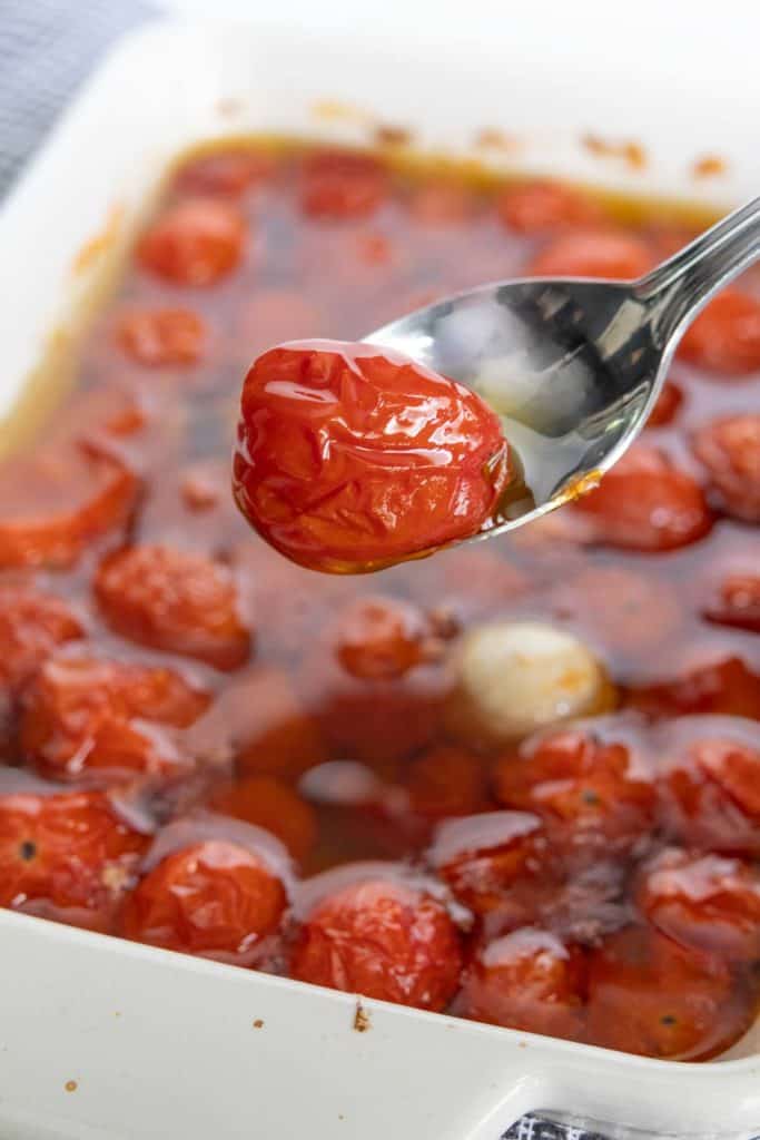 A spoon holding a roasted cherry tomato above a baking dish filled with more roasted cherry tomatoes in oil and herbs.