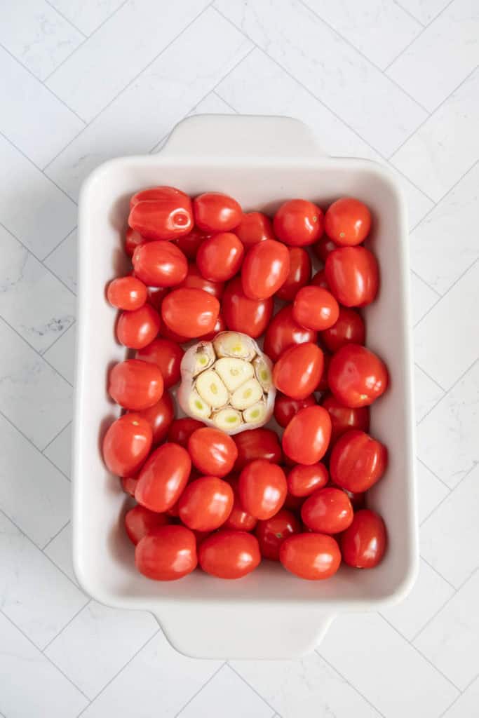 A white rectangular dish filled with numerous small red tomatoes and a single whole garlic bulb in the center.