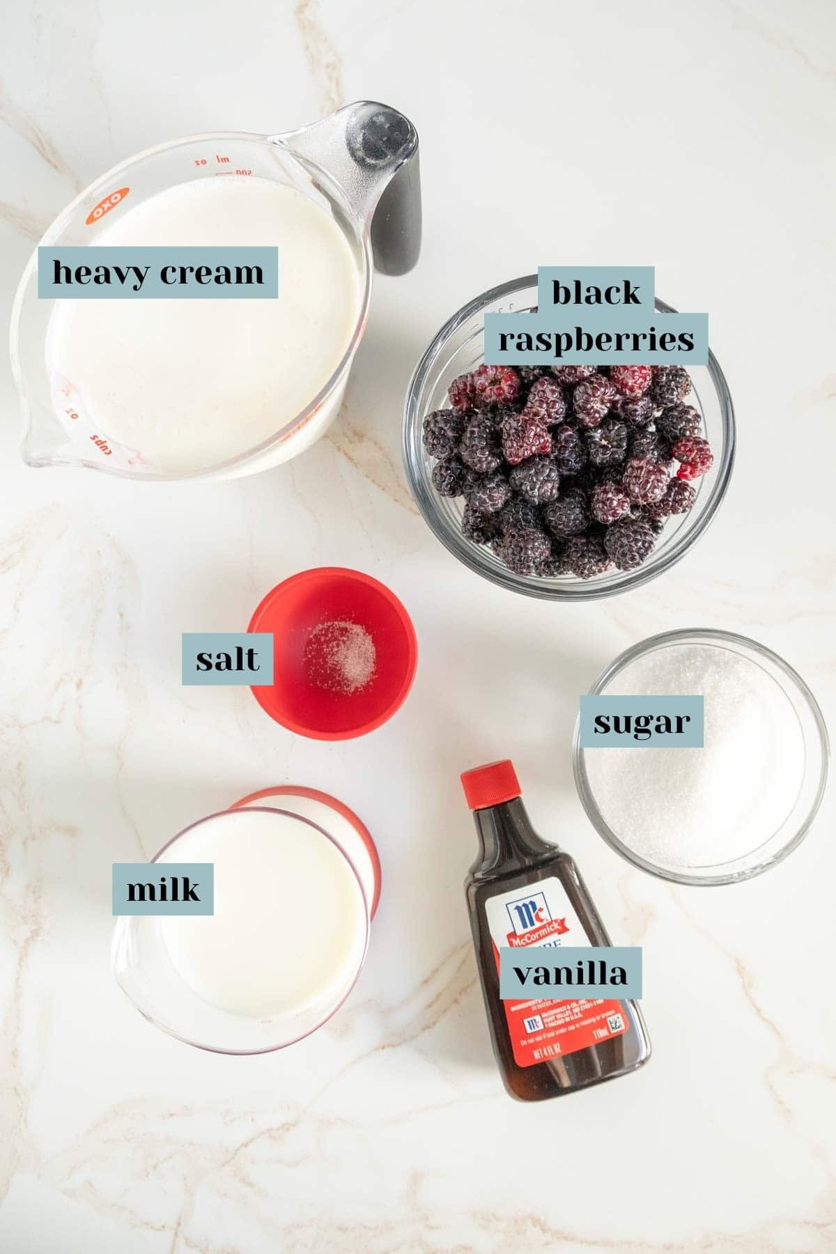 Ingredients laid out on a white surface: a measuring cup of heavy cream, a bowl of black raspberries, a small bowl of salt, a cup of milk, a container of vanilla extract, and a bowl of sugar.