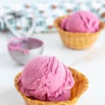 Two scoops of raspberry ice cream served in waffle bowls, with an ice cream scoop and patterned cloth in the background.