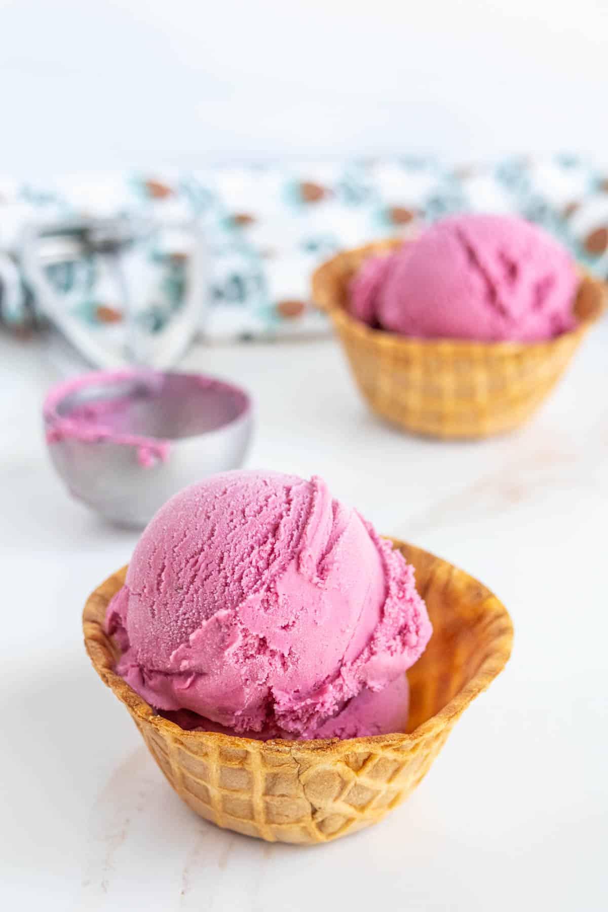 Two scoops of raspberry ice cream served in waffle bowls, with an ice cream scoop and patterned cloth in the background.