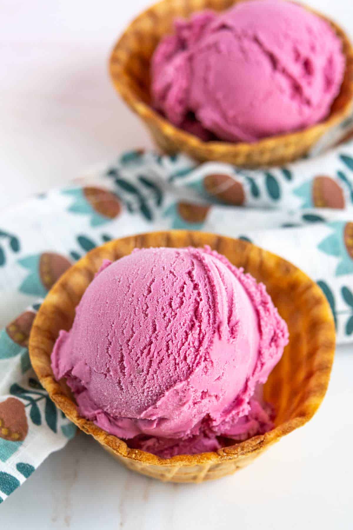 Two scoops of pink ice cream served in waffle bowls, placed on a white surface with a patterned cloth in the background.