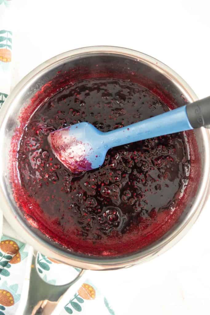 A pot filled with dark berry sauce being stirred with a blue spatula.
