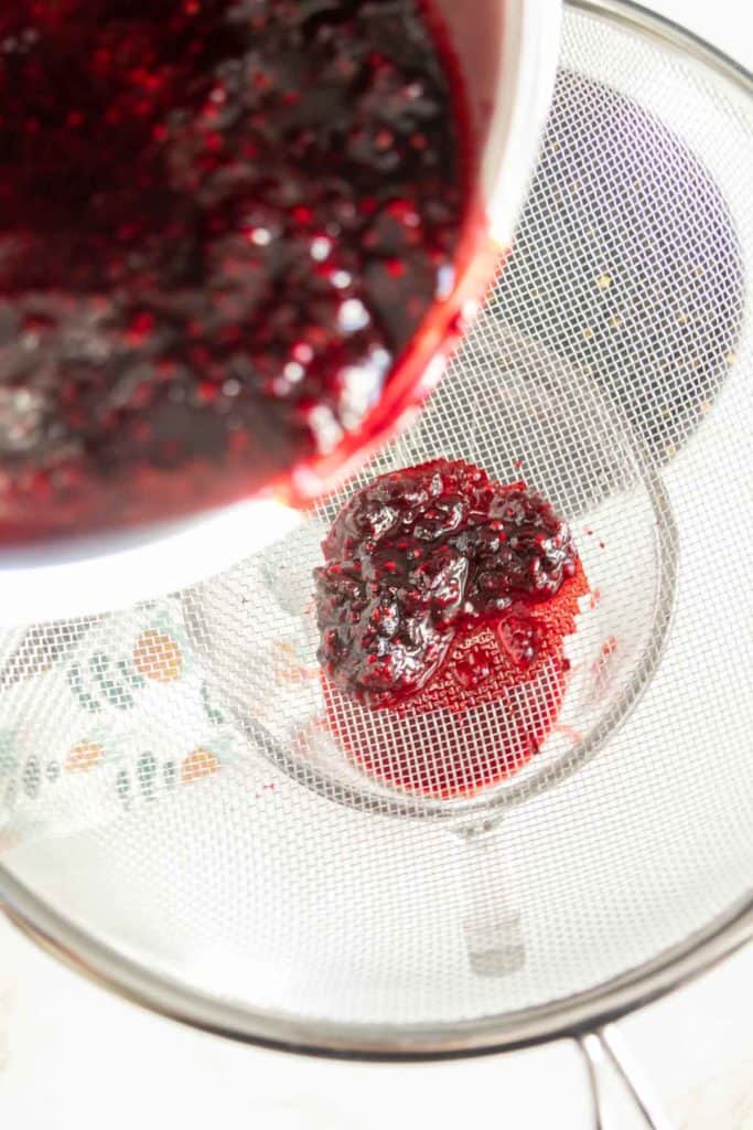 A thick red mixture is being poured through a fine mesh strainer into a container below, separating solids from the liquid.