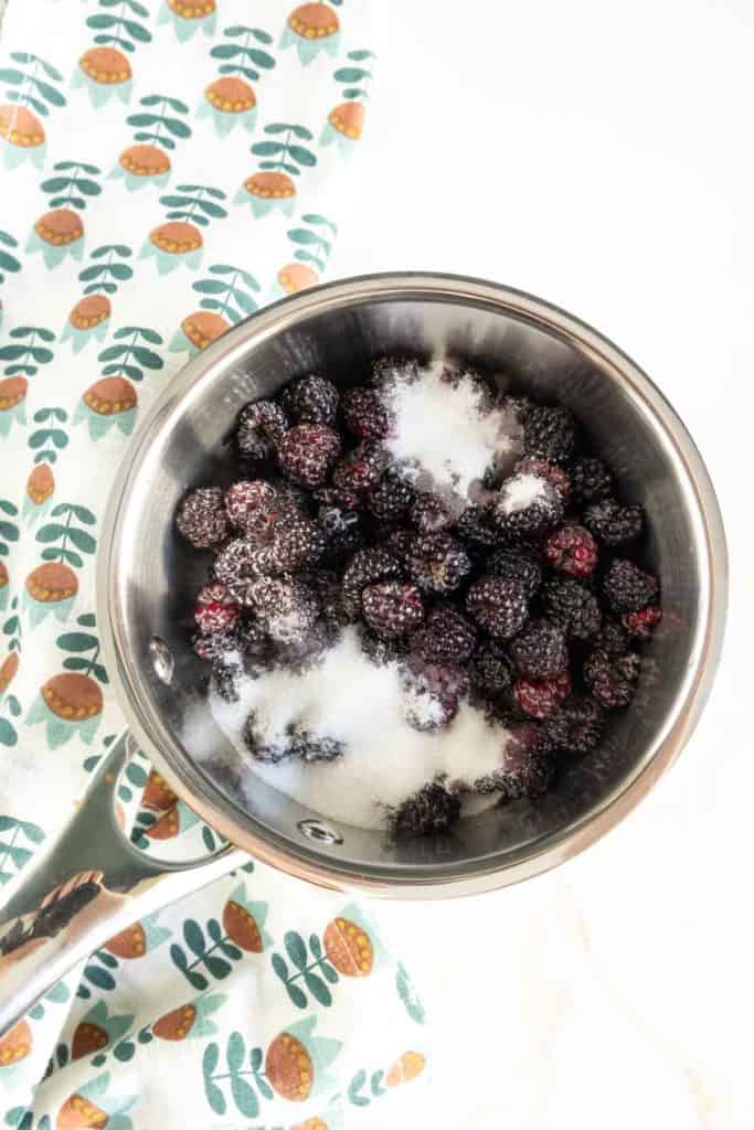 A stainless steel pot filled with blackberries and sugar sits on a white surface next to a patterned cloth.