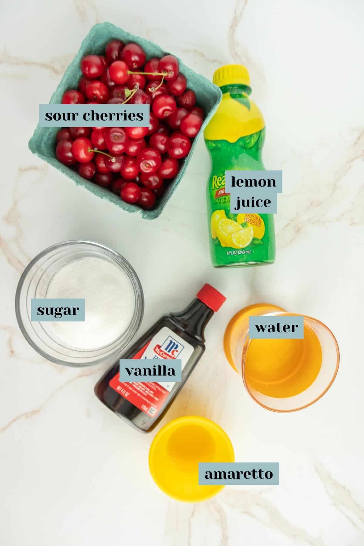 Ingredients for a recipe arranged on a marble surface: a carton of sour cherries, a bottle of lemon juice, a bowl of sugar, a bottle of vanilla extract, a glass of water, and a small bowl of amaretto.