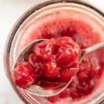 A close-up of a spoonful of bright red cherries being lifted out of a jar filled with cherry compote. A striped towel is visible in the background.