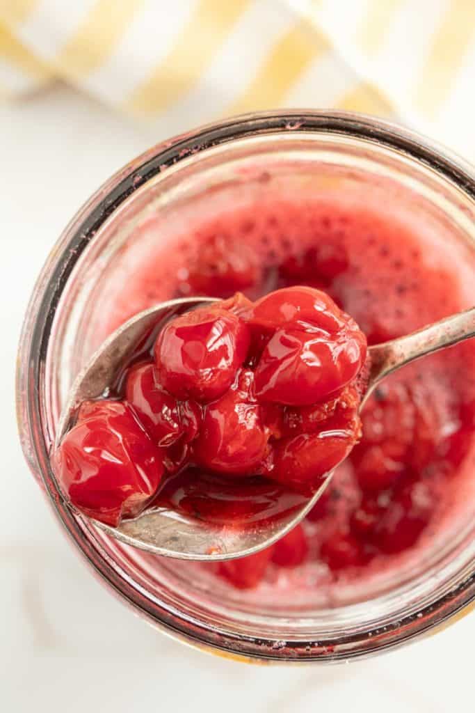 A close-up of a spoonful of bright red cherries being lifted out of a jar filled with cherry compote. A striped towel is visible in the background.