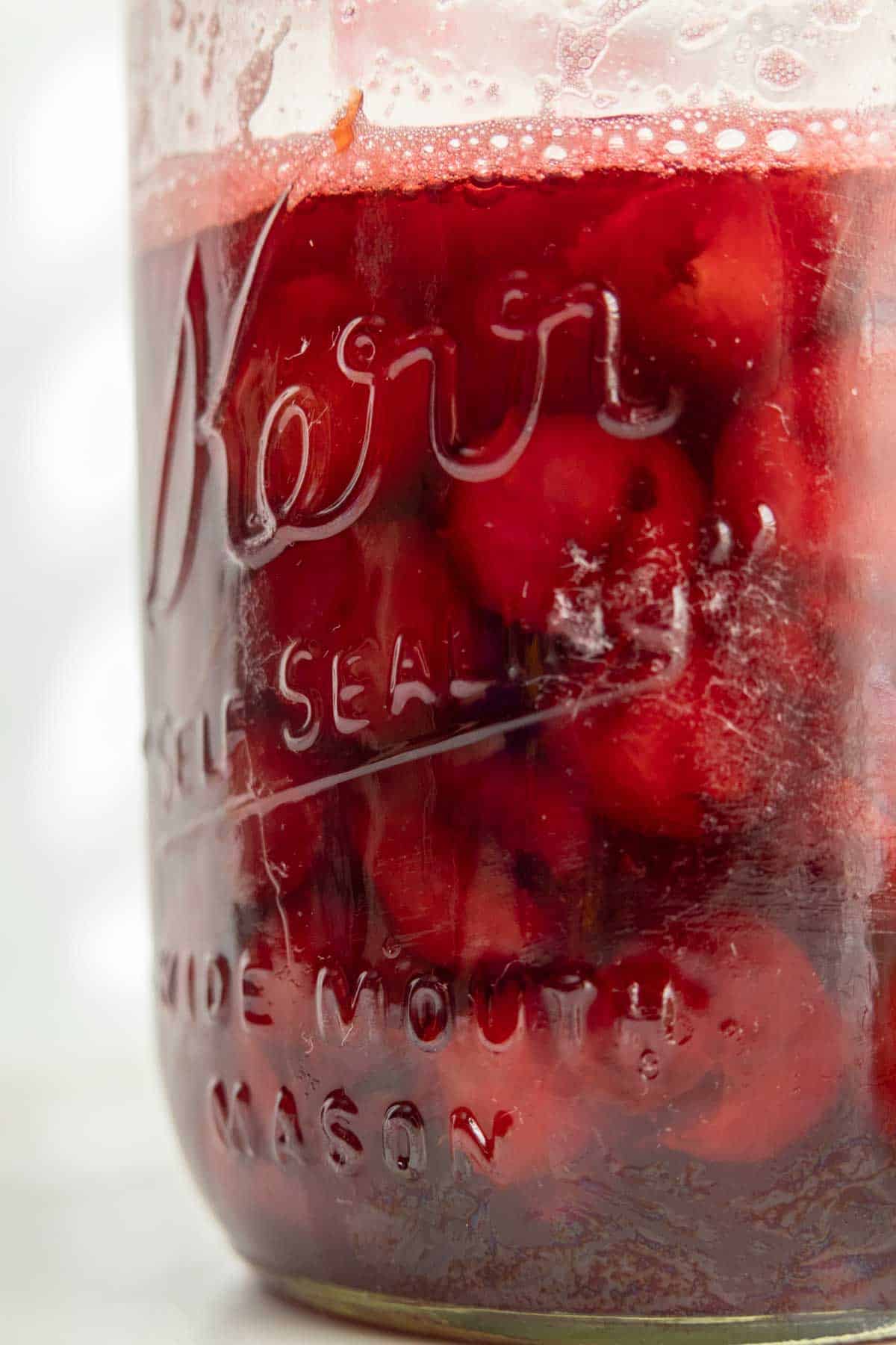 A glass jar filled with red cherries preserved in liquid, with visible bubbles near the top rim. The words "Kerr Self Sealing Wide Mouth Mason" are embossed on the jar's surface.