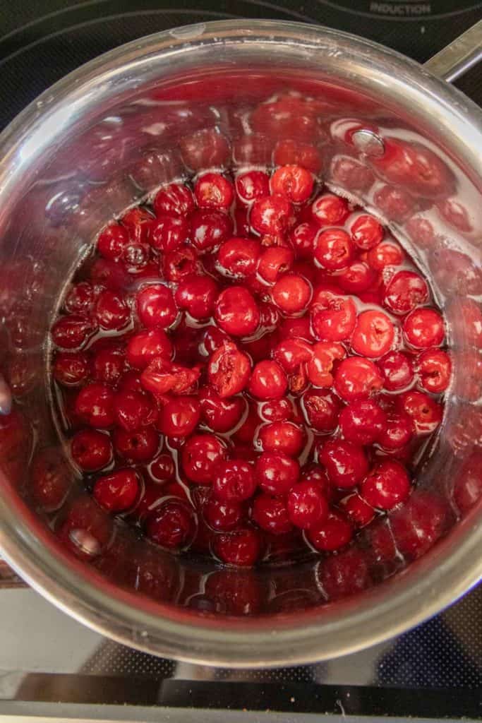 A stainless steel pot filled with bright red cherries in liquid on a stovetop.