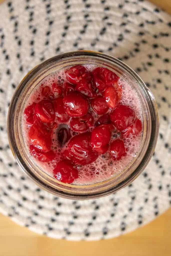 Top view of a jar filled with bright red cherries in liquid, placed on a textured white and black background.