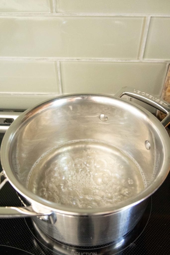 A stainless steel pot filled with boiling water sits on a stovetop. Steam rises from the water as it rapidly bubbles.