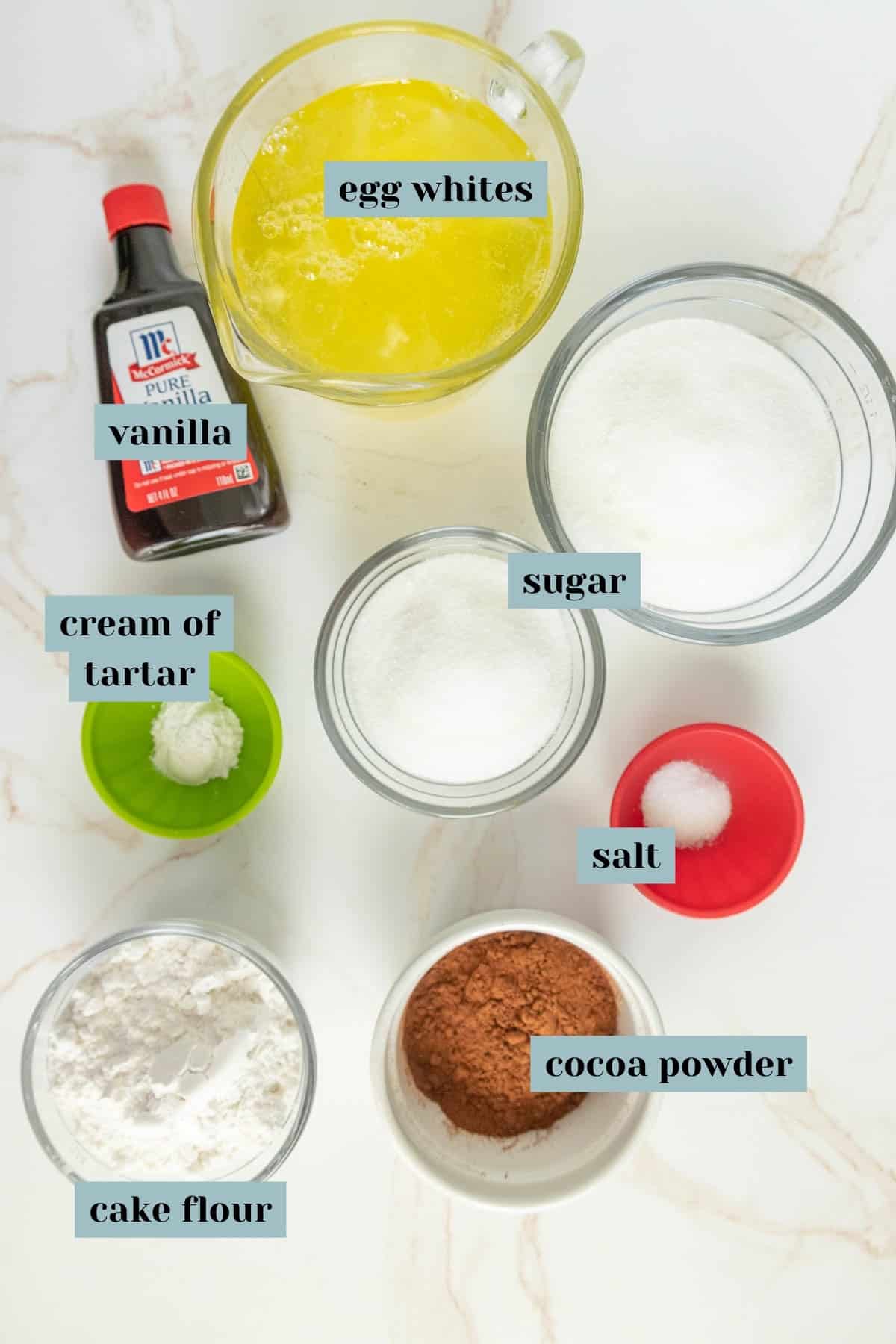 Overhead view of ingredients for baking arranged on a marble surface. Includes egg whites, vanilla extract, sugar, salt, cream of tartar, cocoa powder, and cake flour, each labelled.