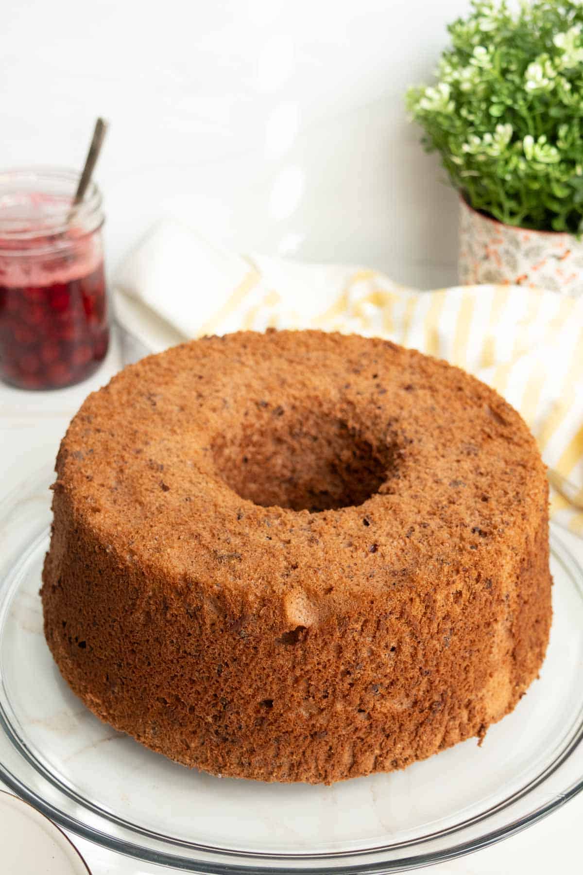 A round, golden-brown Bundt cake sits on a glass plate with a jar of red jam and a green potted plant in the background.