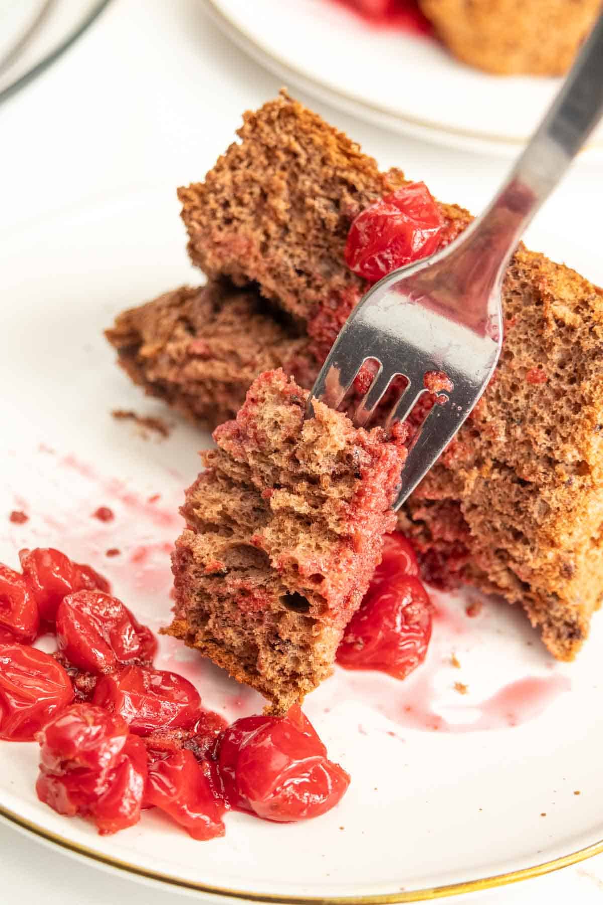 A fork holds a piece of cake with red fruit topping, alongside more fruit and cake pieces on a white plate.