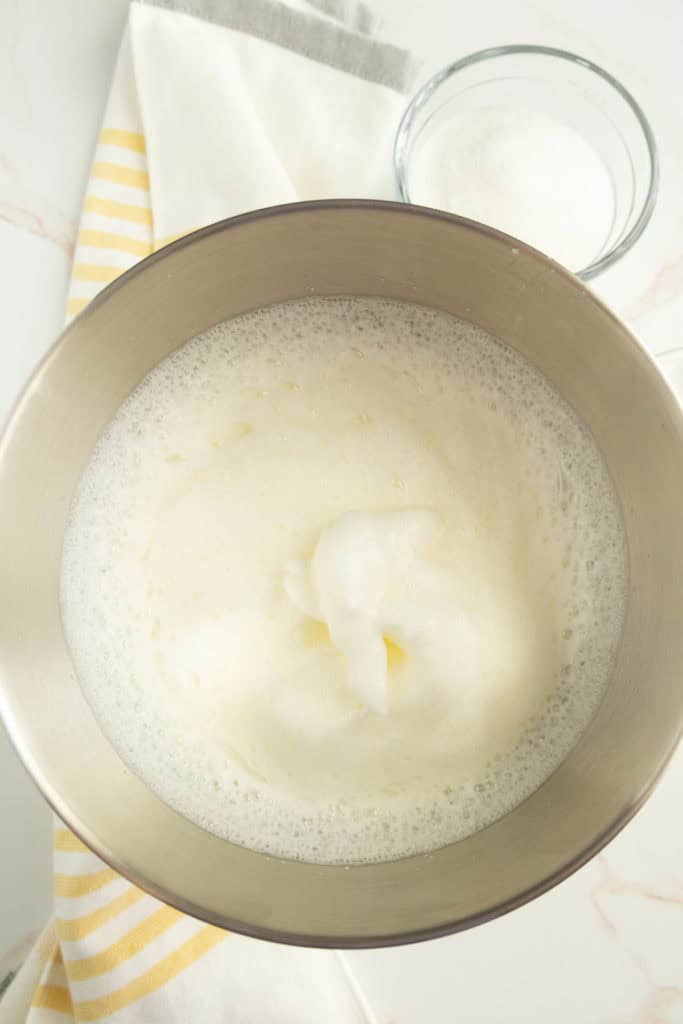 A mixing bowl filled with beaten egg whites is placed on a white surface with a striped yellow and white cloth and a small bowl of sugar nearby.