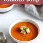 A bowl of Panera tomato soup garnished with croutons and a basil leaf. A spoon and another bowl of soup are in the background. The text "Copycat Panera Tomato Soup" is at the top.