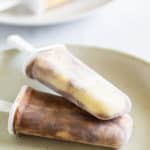 Two swirl-patterned homemade popsicles lie on a beige plate, with more popsicles blurred in the background.