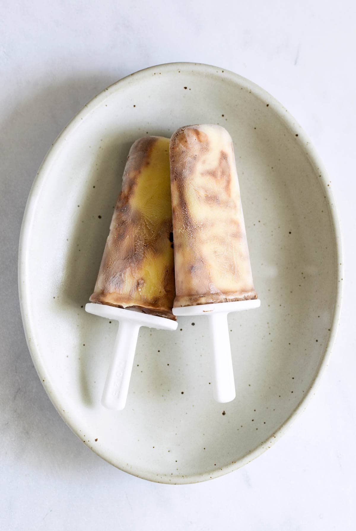 Two popsicles with a marbled pattern of cream and chocolate placed on a white plate.