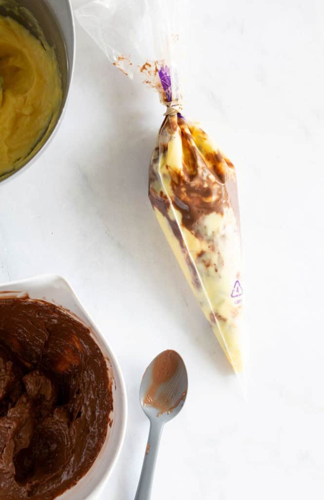 A piping bag filled with a mixture of yellow and brown cream lies next to a bowl of chocolate cream and a spoon on a white surface.