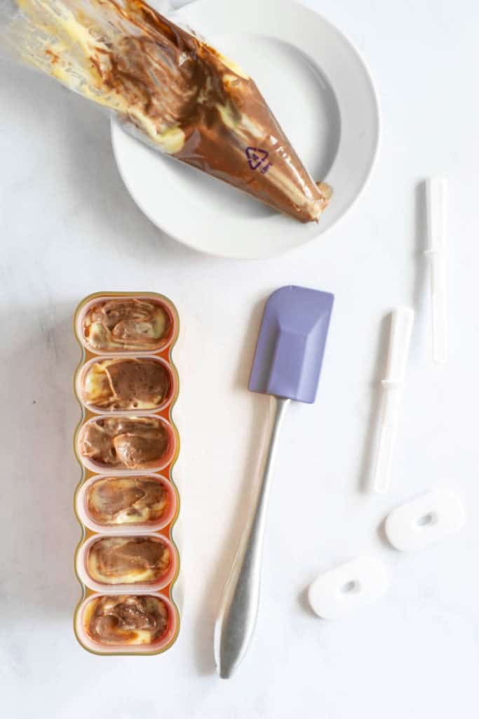 A silicone tray filled with a marbled mixture, likely ice cream or frozen dessert, next to a piping bag, a purple spatula, white lids, and plastic sticks on a white surface.