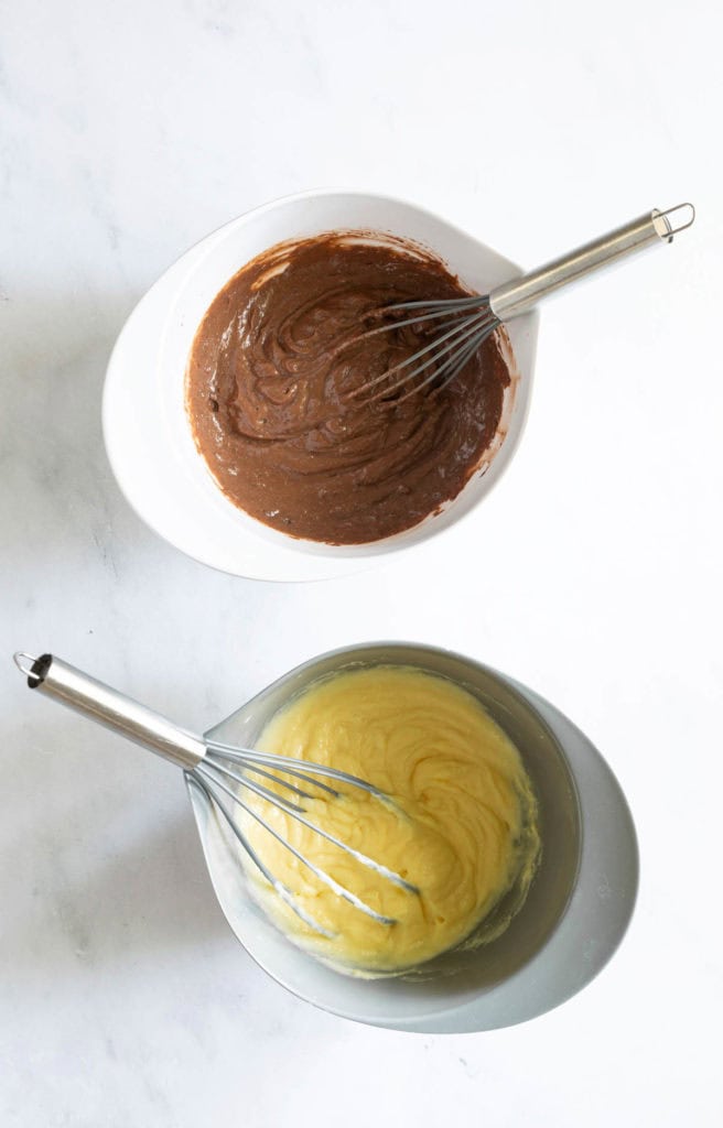 Two mixing bowls: one filled with chocolate batter and the other with yellow batter. Both have a whisk resting inside. The bowls are placed on a light-colored surface.