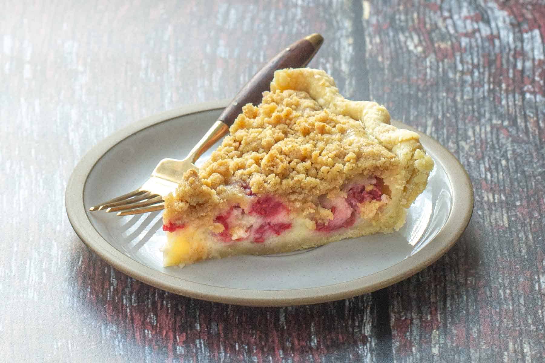 A slice of fruit crumble pie on a plate with a fork, featuring a crumbly top and visible fruit pieces inside.