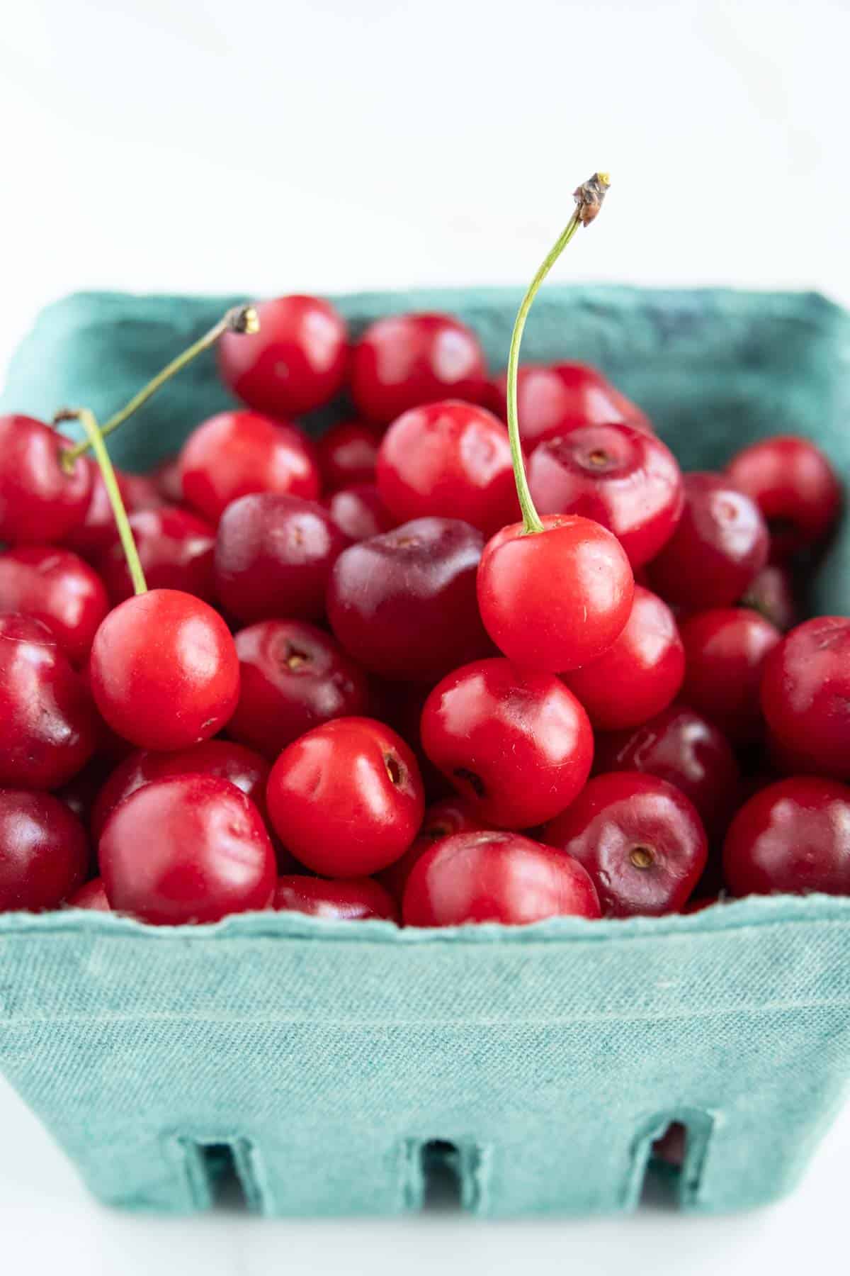 A green cardboard container filled with fresh red cherries, some with stems attached, displayed against a white background.