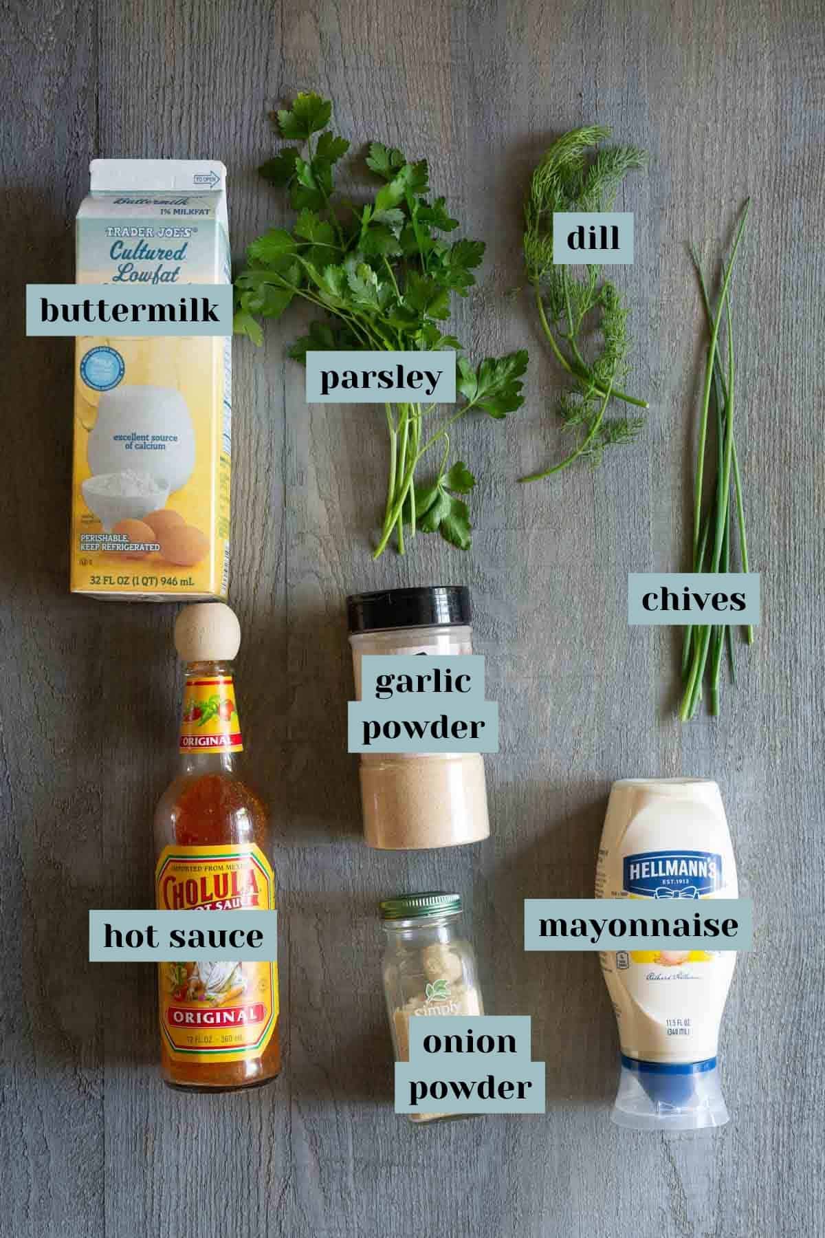 Ingredients for ranch dressing displayed on a wooden surface: buttermilk, parsley, dill, chives, garlic powder, hot sauce, onion powder, mayonnaise. Labels by each ingredient indicate their names.