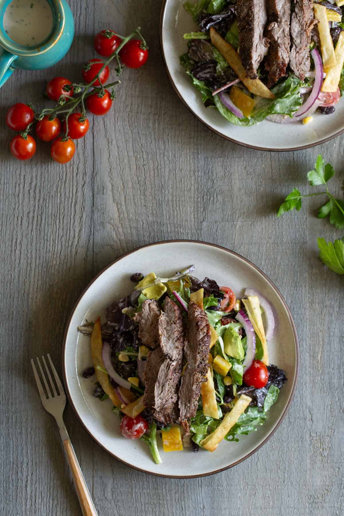 Two plates of steak salad with vegetables, tortilla strips, and dressing on a wooden table, surrounded by a fork, cherry tomatoes on a vine, a sprig of parsley, and a small container of dressing.