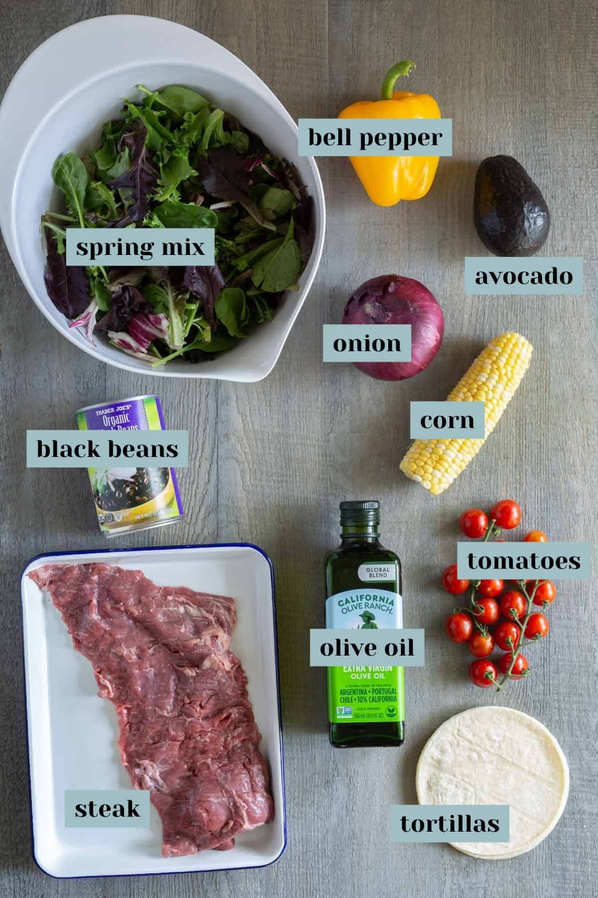 Ingredients for a meal are laid out, including spring mix, bell pepper, avocado, onion, corn, black beans, steak, olive oil, tomatoes, and tortillas on a gray surface.