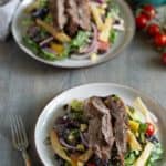 Two plates of steak salad with mixed greens, cherry tomatoes, onion slices, beans, and yellow bell pepper. A pitcher and cherry tomatoes are in the background. Fork placed beside the front plate.