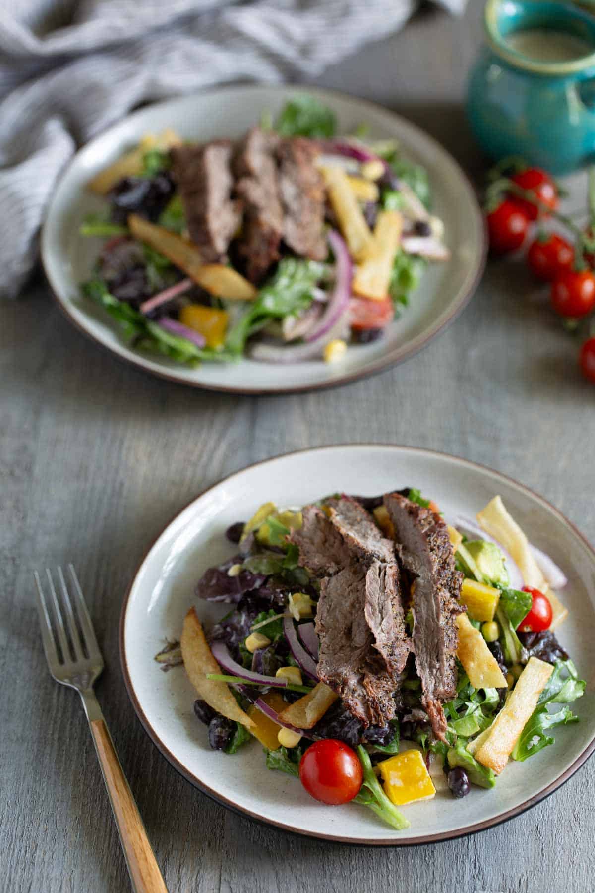 Two plates of steak salad with mixed greens, cherry tomatoes, onion slices, beans, and yellow bell pepper. A pitcher and cherry tomatoes are in the background. Fork placed beside the front plate.