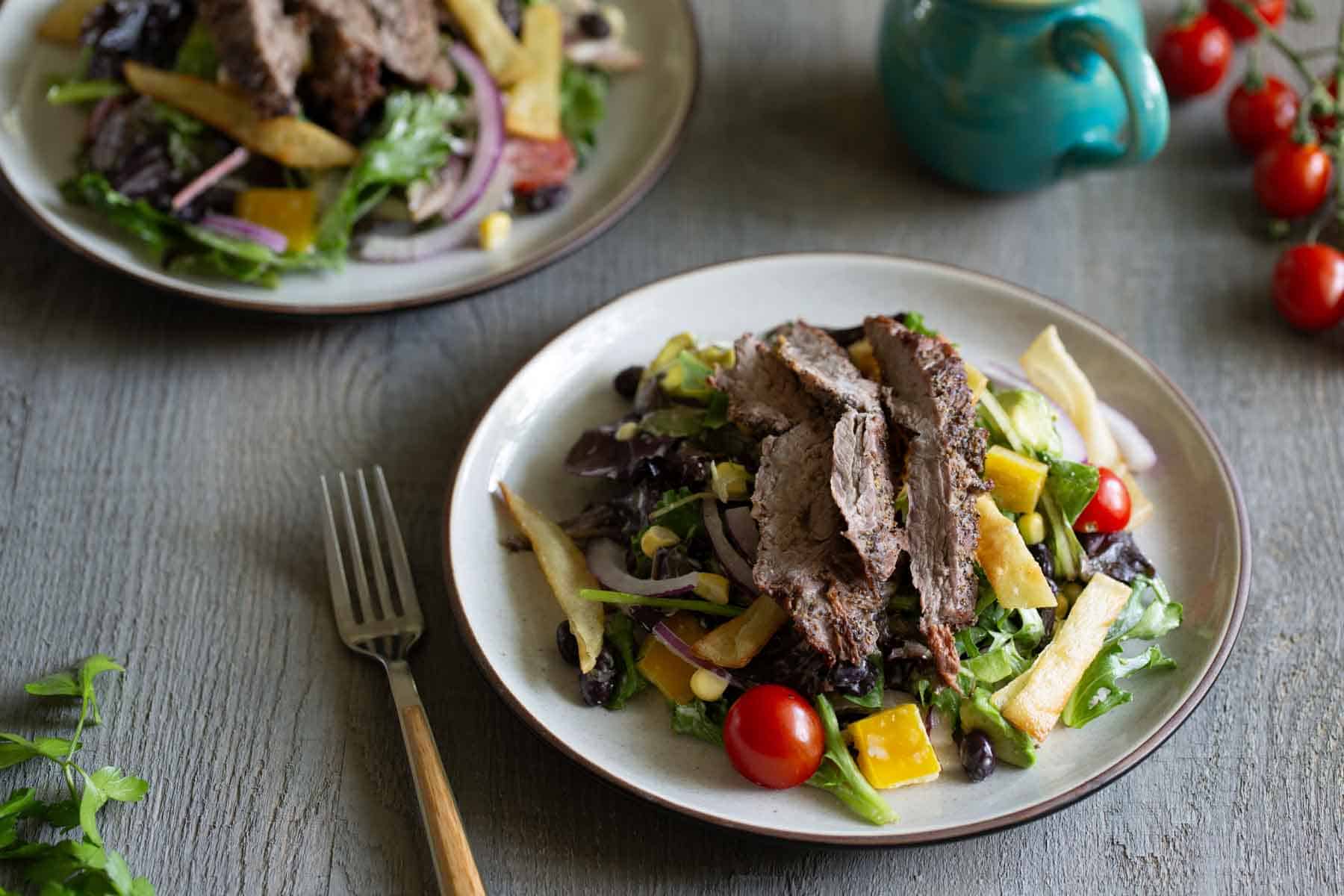 Two plates of salad topped with grilled steak slices and a variety of vegetables, a fork, a turquoise cup, and cherry tomatoes on the side.