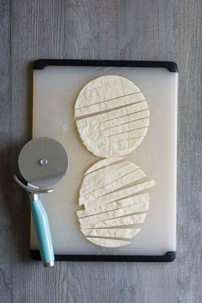 Two round tortillas sliced into strips on a white cutting board with a pizza cutter beside them.