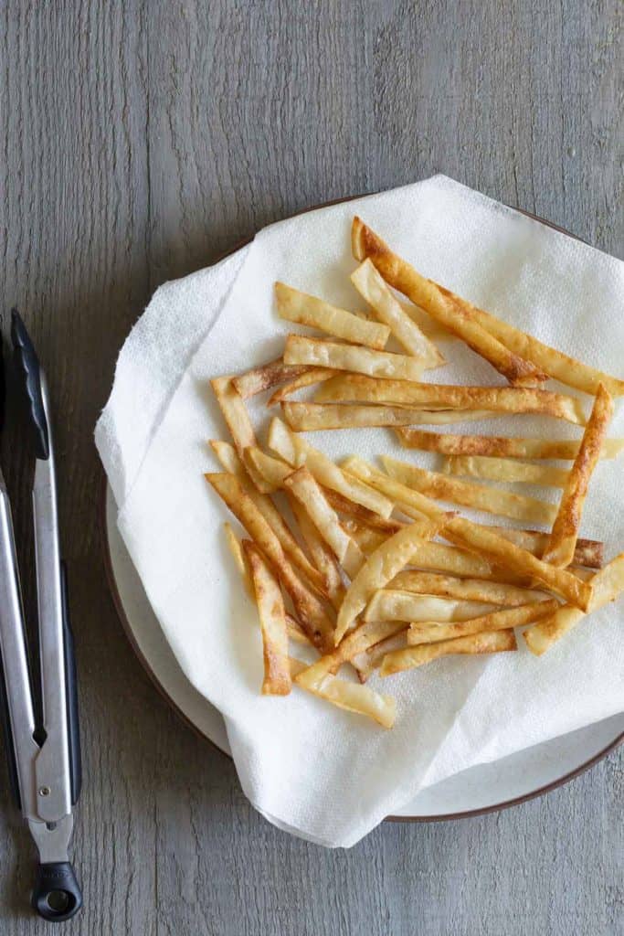 A plate with crispy, golden-brown homemade fries on a paper towel next to a pair of kitchen tongs on a textured gray surface.