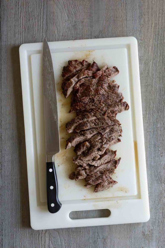 Sliced, cooked meat on a white cutting board, with a large knife placed beside it.