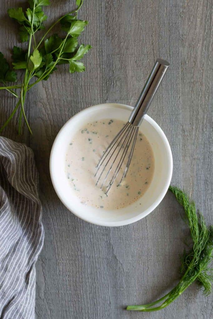 A white bowl filled with a creamy sauce, garnished with herbs and mixed using a whisk. Fresh parsley and dill are placed around the bowl on a wooden surface. A striped cloth is on the left.