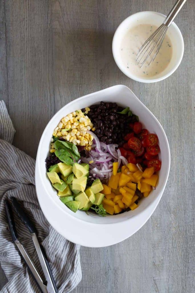 A white bowl contains diced avocado, corn, black beans, cherry tomatoes, red onions, and yellow bell peppers, arranged separately. A bowl of dressing with a whisk and a pair of tongs are nearby.